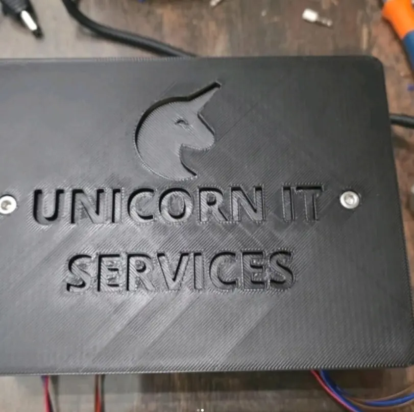 A black 3D-printed plate with the Unicorn IT Services logo and name, screwed onto a hardware build on a workbench